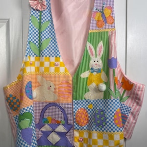 May include: A patchwork Easter vest with a pink satin lining. The vest features various fabric patterns, including gingham and stripes, and is adorned with appliqued Easter-themed designs such as bunnies, eggs, a basket, and a butterfly. The vest is hung on a white hanger.