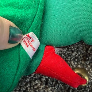 Puede incluir: Primer plano de un gorro de elfo de felpa verde con una punta roja y una campana dorada. El gorro tiene una etiqueta blanca que dice "NEY 20% RAYON CHINA". El gorro est&aacute; sobre una superficie gris texturizada.
