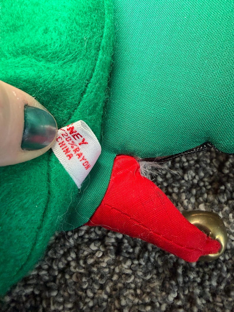 Puede incluir: Primer plano de un gorro de elfo de felpa verde con una punta roja y una campana dorada. El gorro tiene una etiqueta blanca que dice "NEY 20% RAYON CHINA". El gorro est&aacute; sobre una superficie gris texturizada.