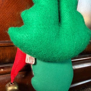 Puede incluir: Adorno navide&ntilde;o vintage de elfo con sombrero y cuerpo de fieltro verde. El elfo tiene un gorro puntiagudo rojo con una campana dorada y una etiqueta que dice "Made in Cotton". El adorno est&aacute; dise&ntilde;ado para colgar.