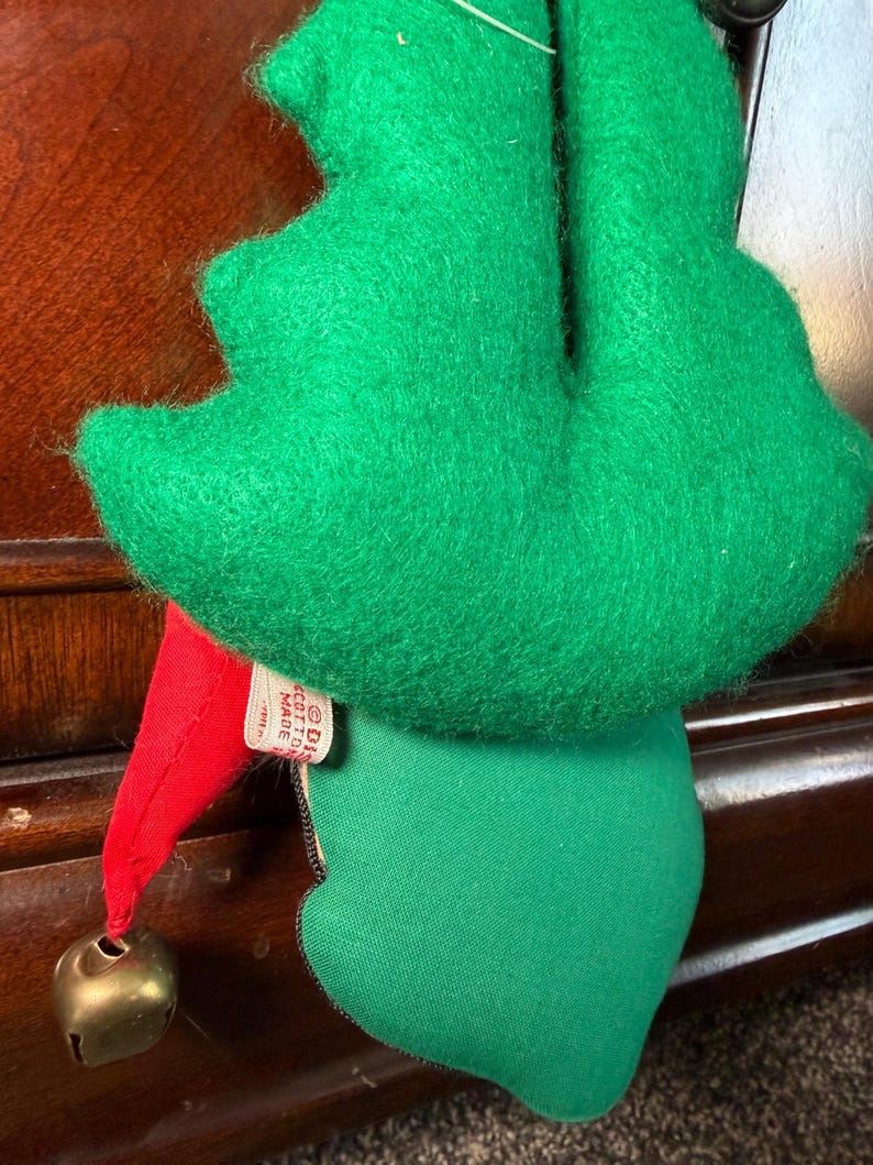 Puede incluir: Adorno navide&ntilde;o vintage de elfo con sombrero y cuerpo de fieltro verde. El elfo tiene un gorro puntiagudo rojo con una campana dorada y una etiqueta que dice "Made in Cotton". El adorno est&aacute; dise&ntilde;ado para colgar.