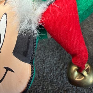 Puede incluir: Primer plano de la cabeza y el sombrero de una mu&ntilde;eca elfo navide&ntilde;a. El elfo tiene una cara bronceada, cabello negro y un sombrero verde y rojo con una campana dorada. El borde blanco y esponjoso acent&uacute;a el sombrero y la cara.