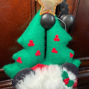 Puede incluir: Un adorno navide&ntilde;o con un &aacute;rbol de fieltro verde con acentos de bayas rojas y una cabeza de Mickey Mouse con un gorro de Pap&aacute; Noel. El adorno tiene una estrella dorada y herrajes decorativos negros.