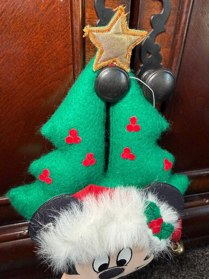 Puede incluir: Un adorno navide&ntilde;o con un &aacute;rbol de fieltro verde con acentos de bayas rojas y una cabeza de Mickey Mouse con un gorro de Pap&aacute; Noel. El adorno tiene una estrella dorada y herrajes decorativos negros.