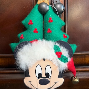 Puede incluir: Adorno navide&ntilde;o para &aacute;rbol con Mickey Mouse. El &aacute;rbol de fieltro verde tiene detalles de bayas rojas y una estrella dorada. Mickey Mouse lleva un gorro de Pap&aacute; Noel con una campana. El adorno est&aacute; dise&ntilde;ado para colgar.