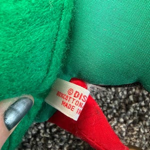 Puede incluir: Primer plano de un gorro de elfo navide&ntilde;o de felpa verde con una punta roja y una campana dorada. El gorro tiene una etiqueta que dice "&copy; DIS 80% COTTON MADE IN". El gorro de elfo est&aacute; hecho de material suave y afelpado.