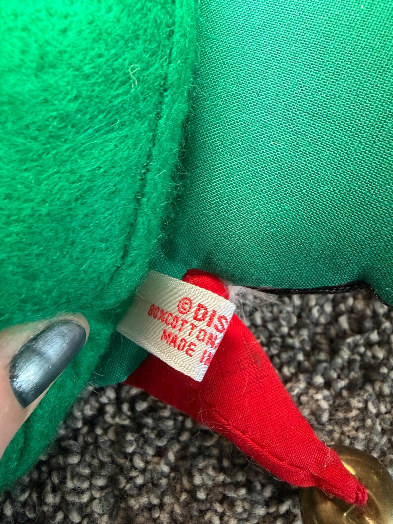 Puede incluir: Primer plano de un gorro de elfo navide&ntilde;o de felpa verde con una punta roja y una campana dorada. El gorro tiene una etiqueta que dice "&copy; DIS 80% COTTON MADE IN". El gorro de elfo est&aacute; hecho de material suave y afelpado.