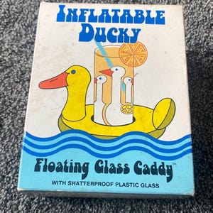 May include: A box for an inflatable floating glass caddy. The box features a yellow duck with an orange beak and a blue eye. The caddy holds three white duck-shaped glasses in a drink with an orange slice.