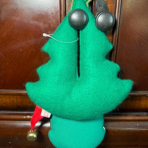Puede incluir: Adorno de &aacute;rbol de Navidad de fieltro verde con grandes ojos de bot&oacute;n negros y una estrella dorada en la parte superior. El adorno tiene un gorro de elfo rojo con una campana dorada. El adorno cuelga de un gancho negro.
