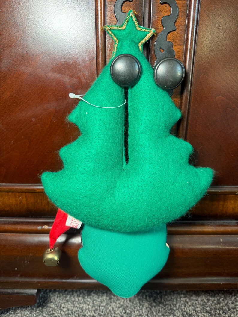 Puede incluir: Adorno de &aacute;rbol de Navidad de fieltro verde con grandes ojos de bot&oacute;n negros y una estrella dorada en la parte superior. El adorno tiene un gorro de elfo rojo con una campana dorada. El adorno cuelga de un gancho negro.