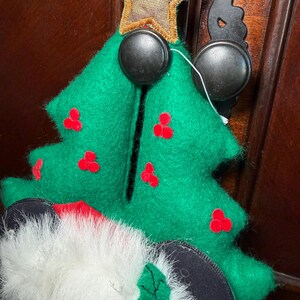 Puede incluir: Adorno de &aacute;rbol de Navidad de fieltro verde con detalles de bayas rojas y una estrella dorada. El adorno est&aacute; sujeto a una superficie de madera marr&oacute;n oscuro con un gancho de metal negro y una cabeza de Mickey Mouse.