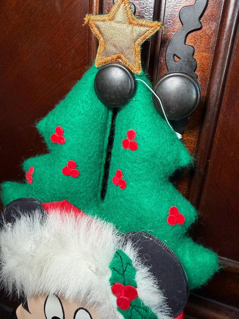 Puede incluir: Adorno de &aacute;rbol de Navidad de fieltro verde con detalles de bayas rojas y una estrella dorada. El adorno est&aacute; sujeto a una superficie de madera marr&oacute;n oscuro con un gancho de metal negro y una cabeza de Mickey Mouse.
