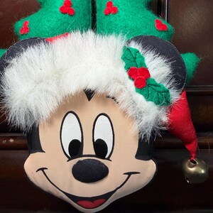 Puede incluir: Una decoraci&oacute;n navide&ntilde;a de Mickey Mouse. La decoraci&oacute;n de felpa presenta la cara de Mickey con un gorro de Pap&aacute; Noel y un &aacute;rbol de Navidad verde. El sombrero tiene un ribete de piel sint&eacute;tica blanca y un cono rojo con una campana dorada. El &aacute;rbol es verde con acentos de bayas rojas.