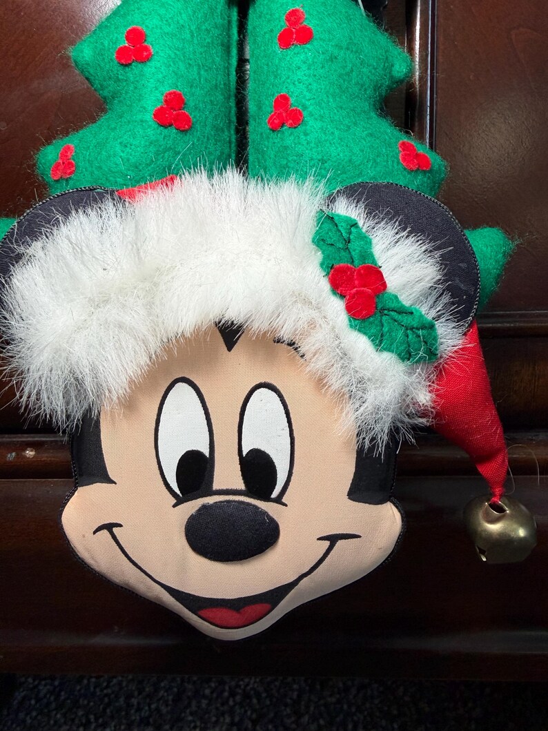 Puede incluir: Una decoraci&oacute;n navide&ntilde;a de Mickey Mouse. La decoraci&oacute;n de felpa presenta la cara de Mickey con un gorro de Pap&aacute; Noel y un &aacute;rbol de Navidad verde. El sombrero tiene un ribete de piel sint&eacute;tica blanca y un cono rojo con una campana dorada. El &aacute;rbol es verde con acentos de bayas rojas.