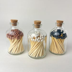 May include: Three clear glass bottles with cork stoppers, each filled with wooden matches. The left bottle contains brown-tipped matches, the center bottle holds white-tipped matches, and the right bottle features black-tipped matches.