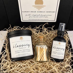 May include: A black gift box containing a black candle jar, a small glass bottle with wooden matches, and a black spray bottle. Each item features a white label with the text Blessing, Black Tea, Toasted Fig, Nutmeg, and Vanilla Tonka.