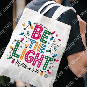 Puede incluir: Una bolsa de lona blanca con las palabras "BE THE LIGHT" en letras coloridas, rodeada por una cadena de luces navideñas. El texto "Matthew 5:14" está impreso debajo. La bolsa tiene correas blancas y se lleva sobre el hombro de una persona.