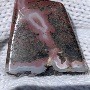 May include: A polished, trapezoidal-shaped agate cabochon featuring a complex pattern of deep red, earthy brown, and black inclusions, accented by concentric bands of white and translucent gray mineral layers throughout the stone surface.
