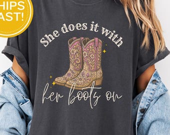 May include: Dark gray t-shirt with the text "She does it with her boots on" and an illustration of cowboy boots. The boots are tan with pink floral accents. A yellow circle with "Ships Fast!" is in the upper left corner.