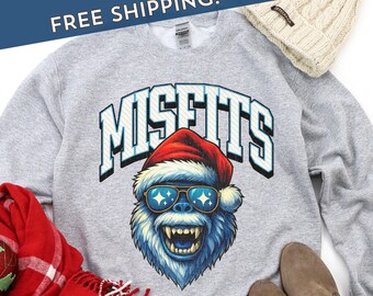 May include: A light gray sweatshirt featuring the word "MISFITS" in a blue and white collegiate font. Below the text is a blue yeti wearing a Santa hat and sunglasses with star-shaped reflections. A red scarf and cream-colored beanie are in the background.
