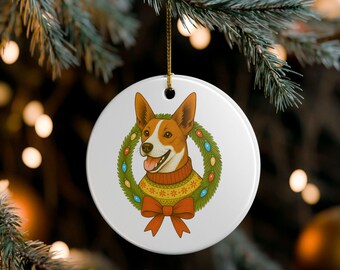 May include: A round white Christmas ornament featuring a dog wearing a sweater and a wreath. The dog has brown and white fur and is smiling. The wreath is green with colorful lights and a red bow.