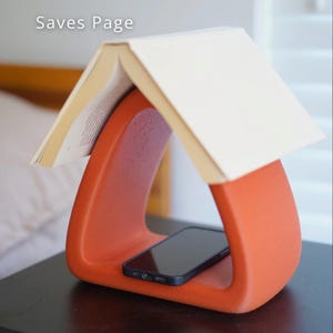 Könnte beinhalten: Ein orangefarbener Buchständer in Dreiecksform mit einem Smartphone im Inneren. Ein Buch liegt obenauf, mit dem Text "Saves Page" sichtbar. Der Ständer besteht aus glattem Material und steht auf einer dunklen Oberfläche.