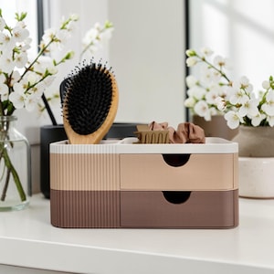 Könnte beinhalten: Ein zweistufiger Organizer in Beige- und Brauntönen mit Fächern. Eine Holzbürste mit schwarzen Borsten und Haargummis liegen obenauf. Weiße Blumen in einer Vase sind im Hintergrund.