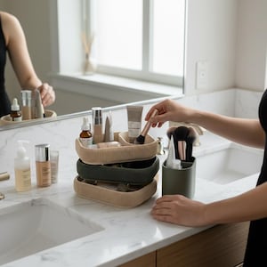 Peut inclure: Trois organisateurs de cosmétiques côtelés beiges et vert foncé, contenant divers produits de beauté et pinceaux de maquillage. Une personne tient un pinceau de maquillage. Les organisateurs sont sur un comptoir blanc dans une salle de bain.