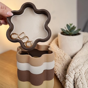 Peut inclure: Un organisateur de bijoux multicouche au design ondulé unique. La couche supérieure, maintenue ouverte, révèle une surface en filet contenant des boucles d'oreilles dorées. L'organisateur est dans les tons de marron, beige et crème, avec une forme de fleur. Une petite plante succulente dans un pot blanc est en arrière-plan.
