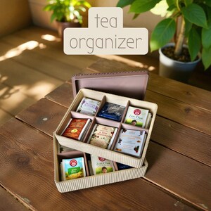 Peut inclure: Un organisateur de thé à deux niveaux avec un extérieur marron clair et des compartiments divisés. L'organisateur est rempli de divers sachets de thé. Le haut de l'organisateur porte les mots "tea organizer" en blanc.