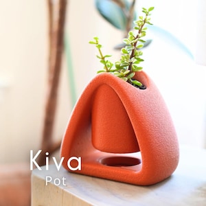 Op de afbeelding: Een oranje Kiva-pot met een uniek, modern ontwerp. De pot heeft een textuur en een opvallende vorm, met een kleinere binnenpot die in een groter frame hangt. Groene plant met kleine bladeren groeit in de pot.