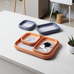Könnte beinhalten: Ein orange-blaues Schreibtisch-Organizer-Set mit drei Tabletts unterschiedlicher Größe. Die Tabletts haben ein geripptes Design und enthalten graue Filzeinlagen. Schlüssel und Büroklammern sind sichtbar. Eine kleine Topf-Sukkulente ist ebenfalls vorhanden.