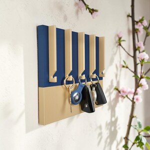 Könnte beinhalten: Ein an der Wand befestigter Schlüsselhalter in Blau und Beige. Der Schlüsselhalter hat beige Haken vor einem blauen Hintergrund. Mehrere Schlüssel hängen an den Haken. Der untere Bereich hat ein strukturiertes, geripptes Design.