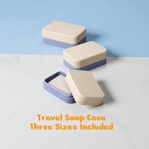 Könnte beinhalten: Drei beige-blaue Reise-Seifenkästchen werden gezeigt, eines davon ist geöffnet und enthält ein Stück Seife. Die Kästchen haben ein geripptes Design auf der blauen Basis und eine glatte Oberseite. Der Text "Travel Soap Case Three Sizes Included" ist sichtbar.
