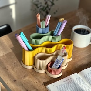 Könnte beinhalten: Ein mehrfarbiger Schreibtisch-Organizer mit wellenförmigem Design, der Stifte und Marker enthält. Der Organizer ist in Gelb-, Grün-, Beige- und Rosatönen gehalten. Eine Tasse Kaffee und ein offenes Buch befinden sich ebenfalls auf der Holzoberfläche.