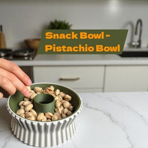 Op de afbeelding: Een groene en witte snackkom gevuld met pistachenoten. De kom heeft een centraal compartiment en een geribbelde, decoratieve buitenkant. De tekst "Snack Bowl - Pistachio Bowl" wordt weergegeven in een groene en oranje banner.