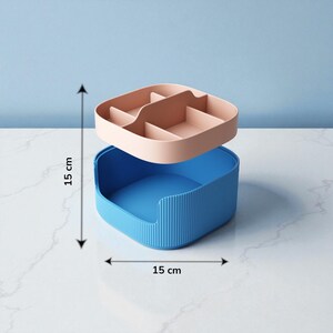 May include: A two-piece storage organizer. The bottom section is blue with vertical ridges, and the top section is divided into compartments and is a light pink color. The organizer is 15 cm wide and 15 cm tall.
