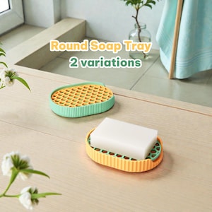Peut inclure: Deux porte-savons ronds avec une surface à motif en nid d'abeille et des côtés nervurés. Un plateau est vert clair, l'autre est jaune avec un pain de savon rectangulaire blanc posé dessus. Le texte Round Soap Tray 2 variations est visible.