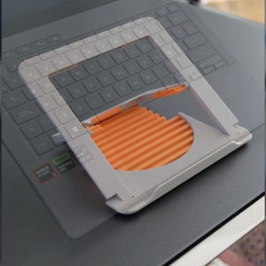 Peut inclure: Un support d'ordinateur portable gris avec une surface orange et striée. Le support est conçu pour maintenir un ordinateur portable incliné. Le support est placé sur un clavier d'ordinateur portable.