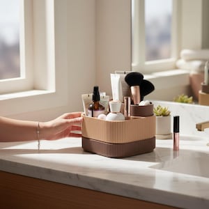 Könnte beinhalten: Ein zweistufiger, geriffelter Kosmetik-Organizer in Beige- und Brauntönen, gefüllt mit Schönheitsprodukten. Der Organizer steht auf einer Marmorarbeitsplatte, eine Hand greift danach. Make-up-Pinsel, Flaschen und Tuben sind sichtbar.