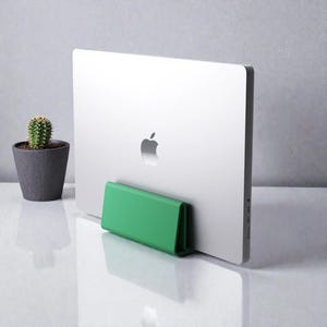 Könnte beinhalten: Ein silberner Laptop mit dem Apple-Logo wird von einem hellgrünen Laptop-Ständer gehalten. Links steht ein kleiner Kaktus in einem grauen Topf. Der Ständer ist leuchtend grün und hat eine gerippte Textur.