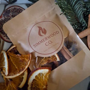 May include: A brown paper bag with a clear window, labeled "Simmerwood Co." contains cinnamon sticks and other spices. Dried orange slices are scattered around the bag. A sprig of evergreen and a decorative tray are in the background.