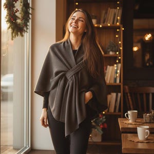 Może przedstawiać: Kobieta w ciemnoszarej dzianinowej pelerynie z unikalnym zapięciem na supeł. Peleryna jest narzucona na czarny top i spodnie. Tło obejmuje okno, półki z książkami i stół z kubkami.