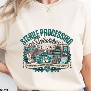May include: An ivory t-shirt featuring a graphic with the text STERILE PROCESSING TECHNICIAN OLIVIA. The design includes medical equipment, a sterilization machine, and a banner with the words DECONTAMINATE INSPECT ASSEMBLE STERILIZE REPEAT.