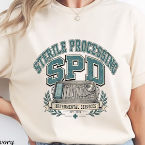 May include: An ivory t-shirt featuring a graphic with the text STERILE PROCESSING SPD and INSTRUMENTAL SERVICES EST. 2026. The design includes a metal tray filled with various surgical instruments, framed by green laurel leaves and a small cross.