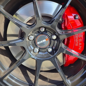 Puede incluir: Una llanta de aleación negra brillante de múltiples radios con una tapa central con el logotipo Nismo. Detrás de los radios, se ve una pinza de freno roja brillante con la palabra NISSAN en blanco sobre un disco de freno plateado.