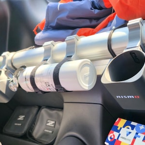 May include: A silver horizontal bar mounted in a vehicle interior features two black cup holders with the red and white nismo logo, and a white cylindrical fire extinguisher secured with black straps. A colourful geometric box sits below.