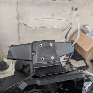 May include: A motorcycle windshield with a clear, curved top section and a black mounting bracket. The bracket has four screw holes and is attached to the motorcycle's dashboard. The windshield is designed to deflect wind.