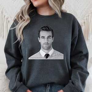 May include: Dark gray sweatshirt featuring a black and white image of James Van Der Beek. The image shows him wearing a white shirt, tie, and sweater. The text "James Van Der Beek" and a signature are below the image.