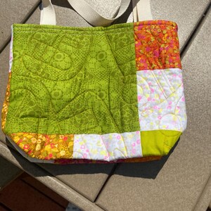 May include: A patchwork tote bag with cream-colored handles. The bag features a quilted design with various fabric patterns, including green, orange, yellow, and white. The bag is sitting on a brown surface.
